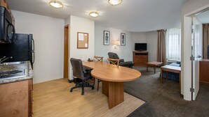 A hotel room with a kitchen area, a dining table with chairs, a TV, and a window with curtains.