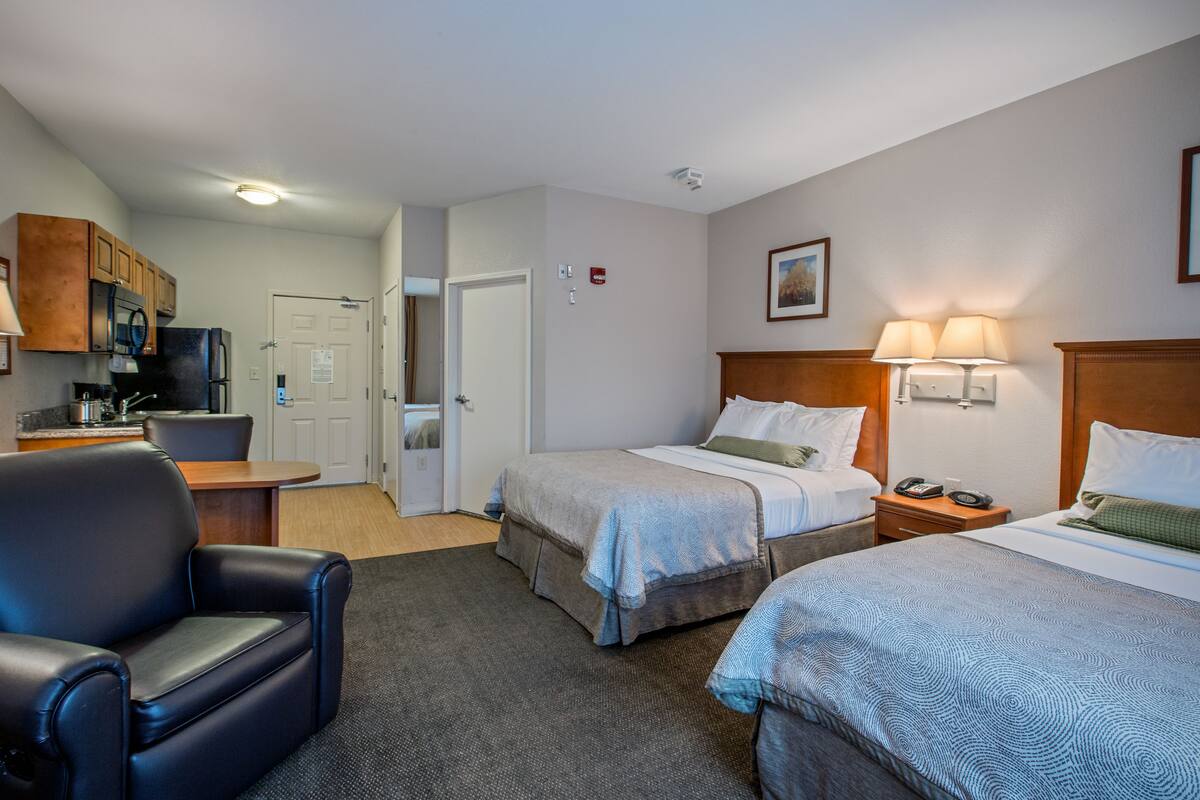 A hotel room with two beds, a desk, a chair, a kitchenette, and a door leading to another room.