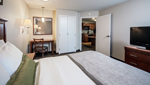 A hotel room with a bed, a desk, a chair, a television, and a kitchen area visible through an open door.