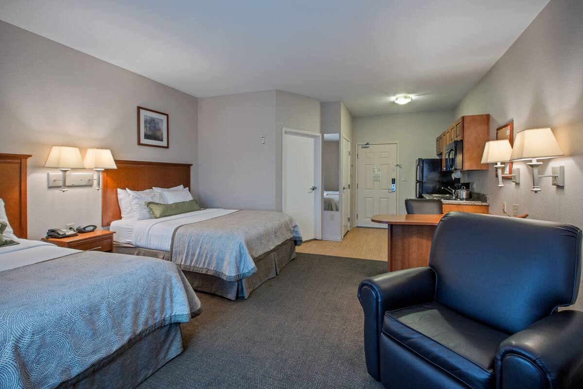 A hotel room with two beds, a desk, a chair, and a kitchenette.