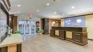 Zona de recepción de un hotel con puerta de vidrio, mostrador de recepción y planta en maceta.