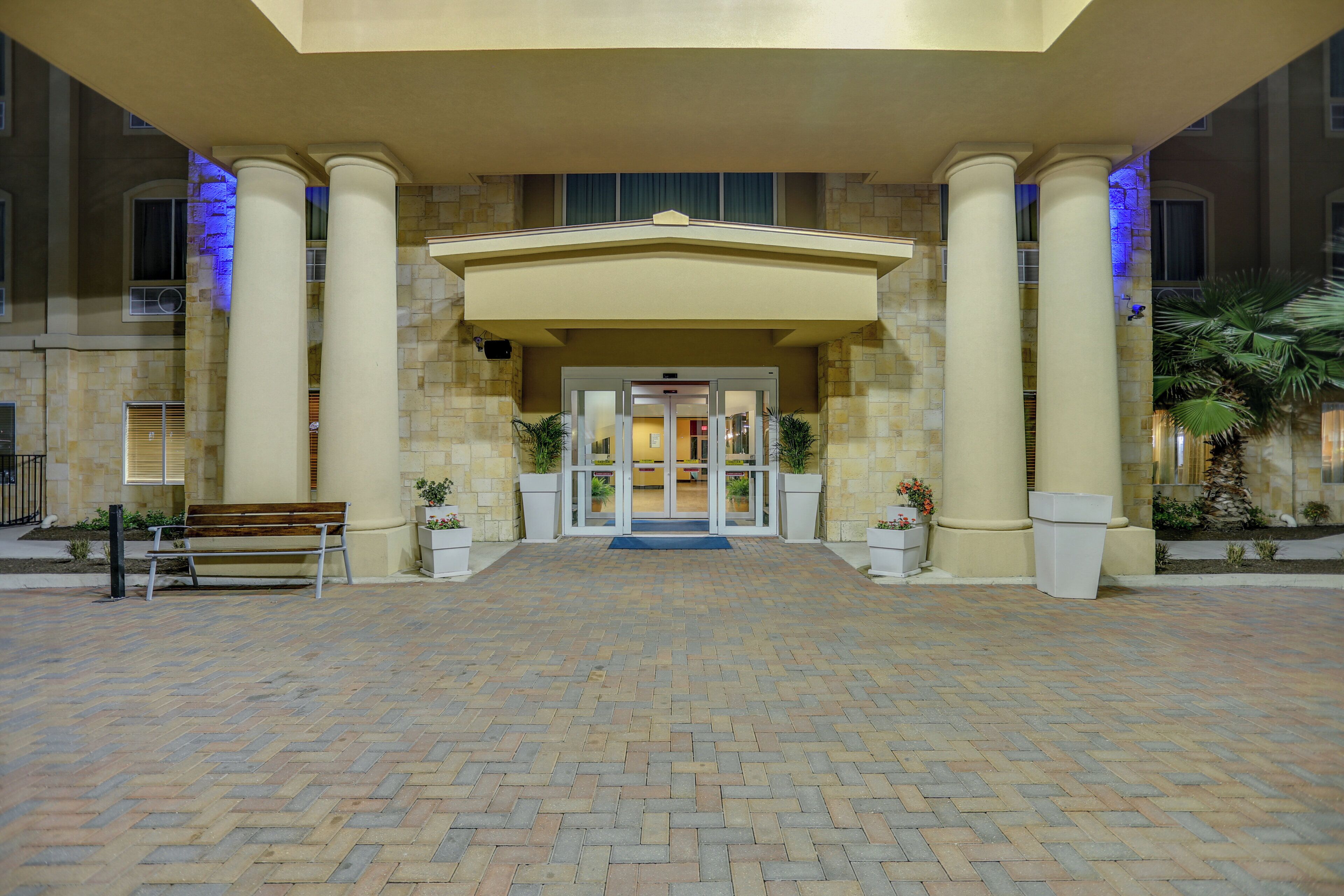 A hotel entrance with a brick-paved driveway, a bench, and a double door entrance.