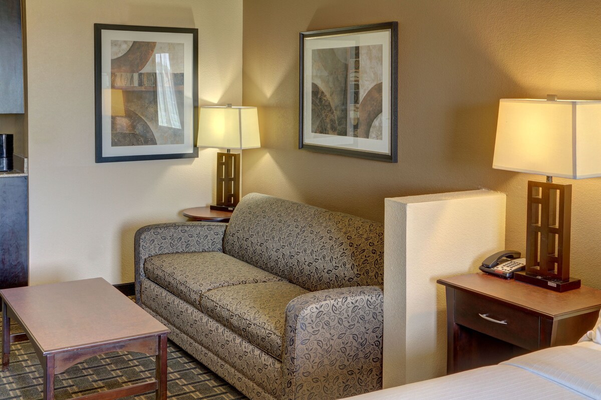 A hotel room with a sofa, a small table, a bedside table with a lamp, a phone, and a framed picture on the wall.