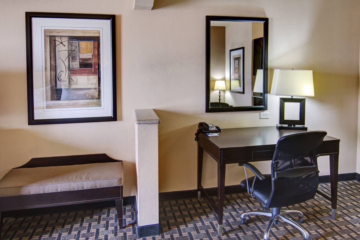 A hotel room with a desk, chair, bedside table, lamp, and a framed abstract artwork.