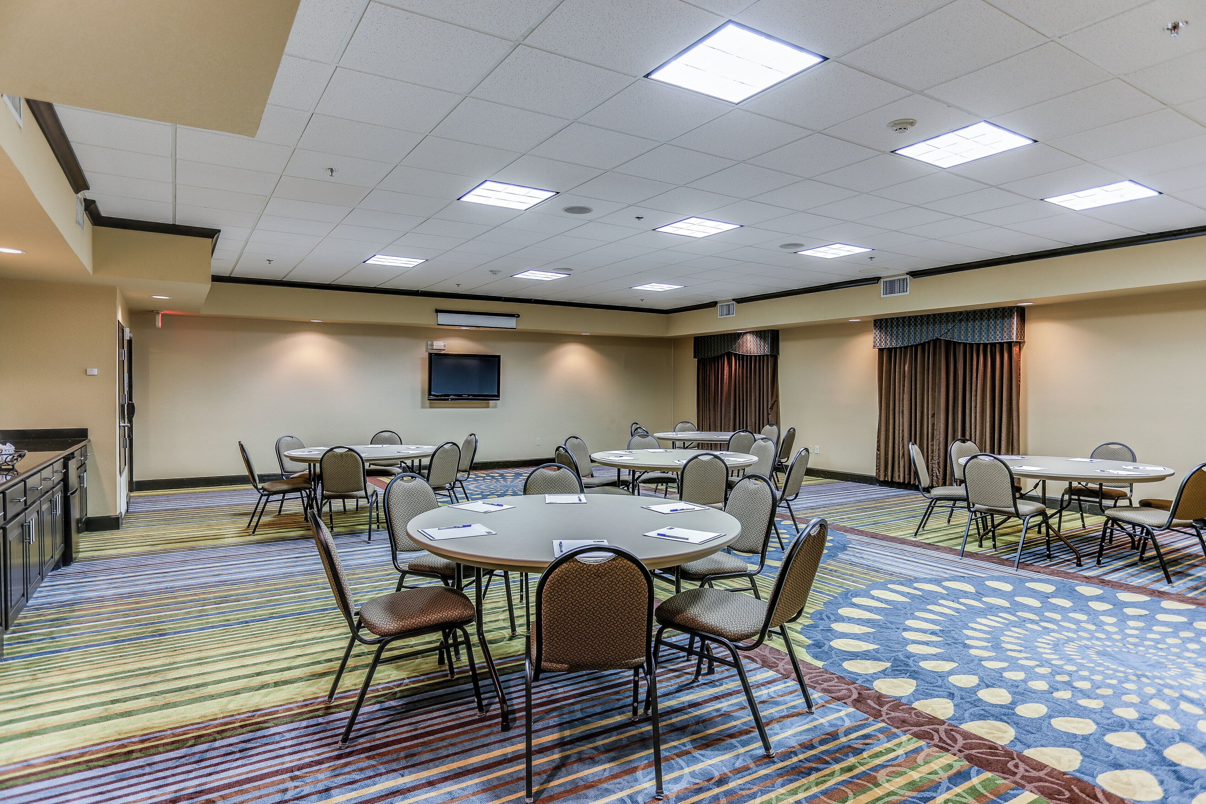 A conference room with multiple tables and chairs, a flat-screen TV, and a patterned carpet.