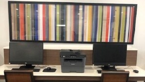 Habitación de hotel con escritorio, dos monitores, una impresora y una obra de arte grande con rayas en la pared.