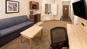 Habitación de hotel con sofá, una mesita, escritorio con silla, microondas, lavabo y televisión.