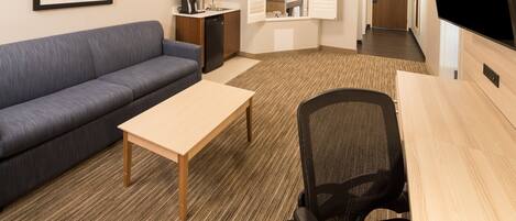 A hotel room with a sofa, a small table, a desk with a chair, a microwave, a sink, and a TV.