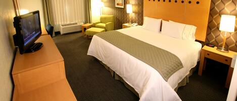 A hotel room with a bed, a desk with a computer, a chair, a bedside table with a lamp, and a wall-mounted TV.