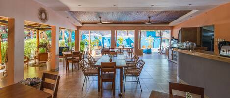 A spacious dining area with multiple tables and chairs, a kitchen area with a counter, and large windows offering views of the outdoors.