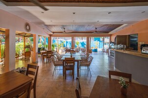 Un comedor amplio con varias mesas y sillas, una zona de cocina con mostrador, y ventanas grandes que ofrecen vistas al exterior.