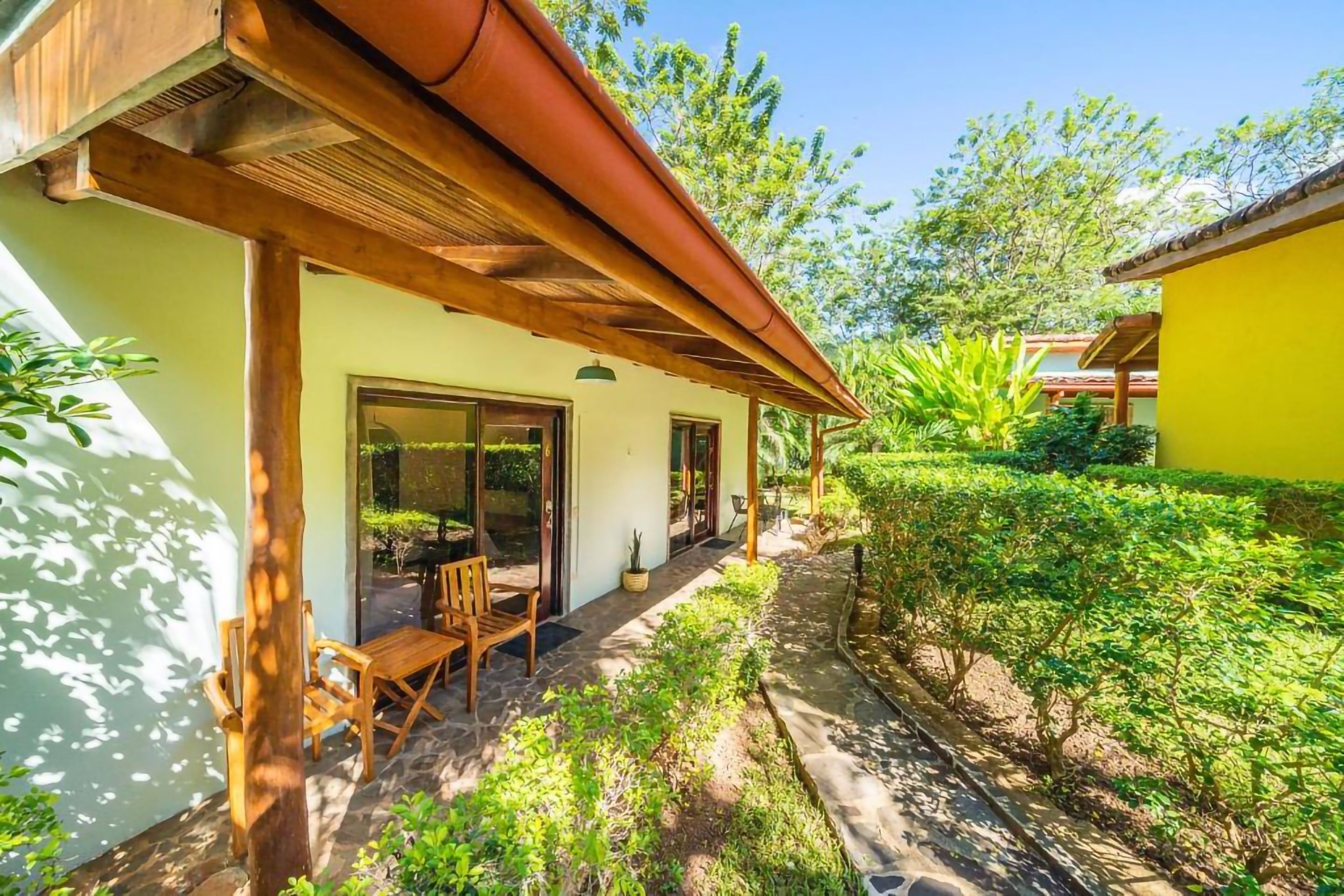 A covered outdoor seating area with wooden furniture, a stone pathway, and lush greenery.