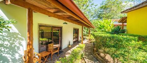 A covered outdoor seating area with wooden furniture, a stone pathway, and lush greenery.