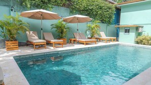 A pool area with wooden lounge chairs and two umbrellas, surrounded by greenery and a tiled deck. - Hotel Arco Iris (Tamarindo)