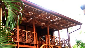 A wooden structure with a staircase leading to a covered area, surrounded by greenery and yellow flowers. - Hotel Arco Iris (Tamarindo)