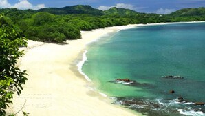 A beach with clear turquoise water, white sand, and lush green vegetation. - Hotel Arco Iris (Tamarindo)