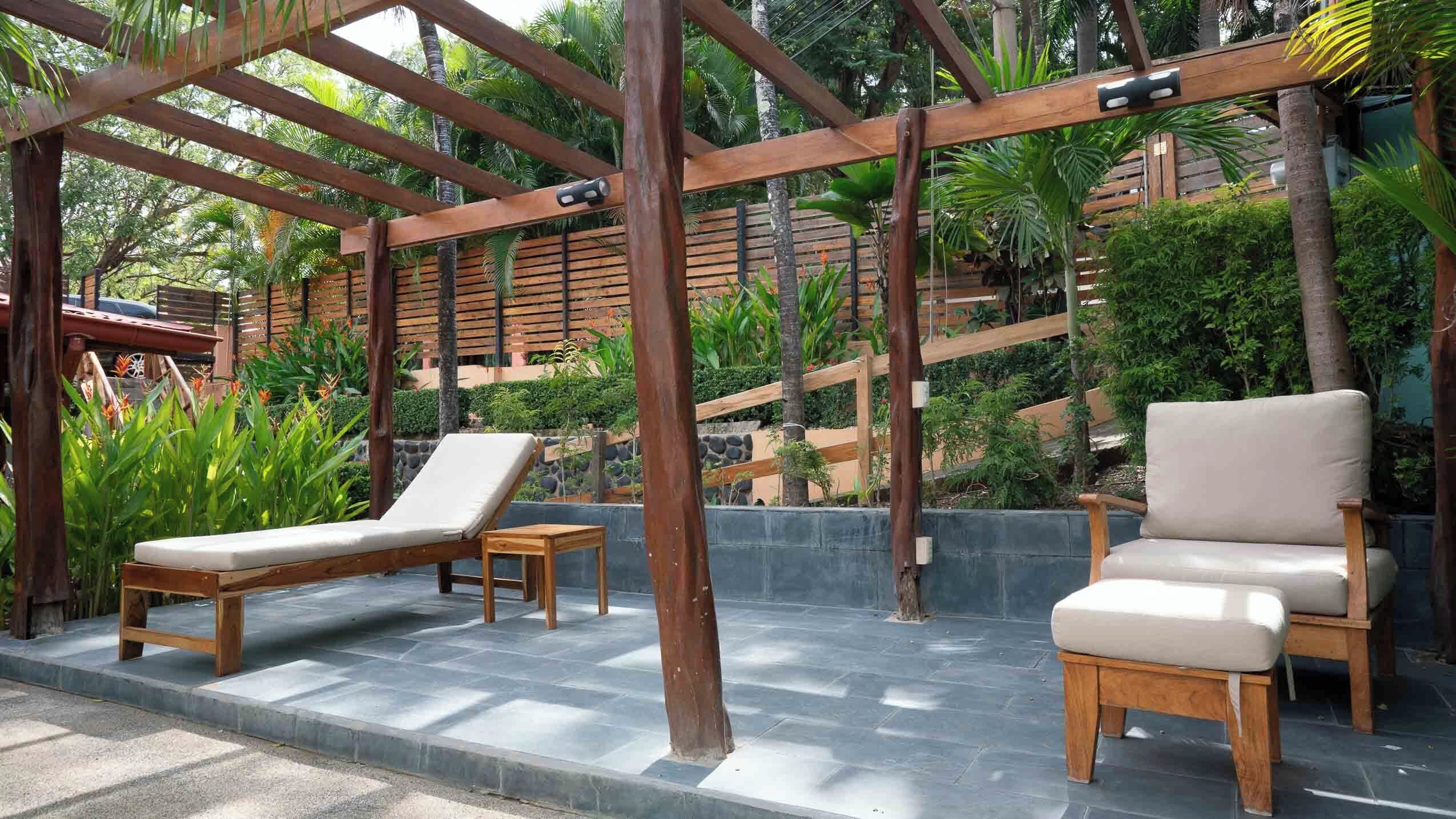 A wooden pergola with two lounge chairs and a small table, surrounded by lush greenery and a stone wall.
