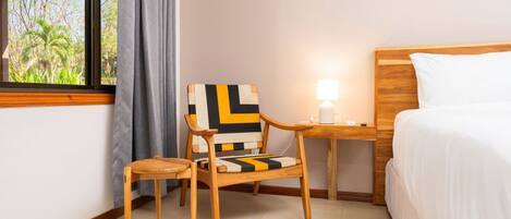 A hotel room with a bed, a chair, a small table with a lamp, and a window with a view of trees.