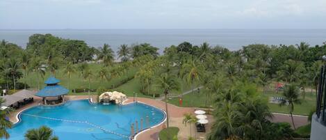 A resort pool with a waterfall feature, surrounded by palm trees and a view of the ocean.