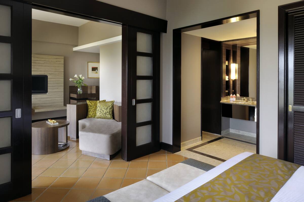 A modern hotel room with a bed, a seating area, a television, and a bathroom with a mirror and sink.