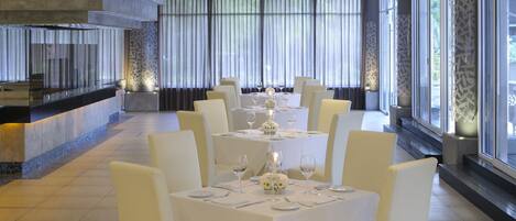 A modern dining area with white tables and chairs, a kitchen counter, and large windows with curtains.