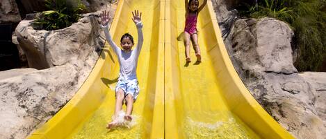 Dos niños resbalando por un tobogán acuático amarillo, con formaciones rocosas y follaje verde de fondo.