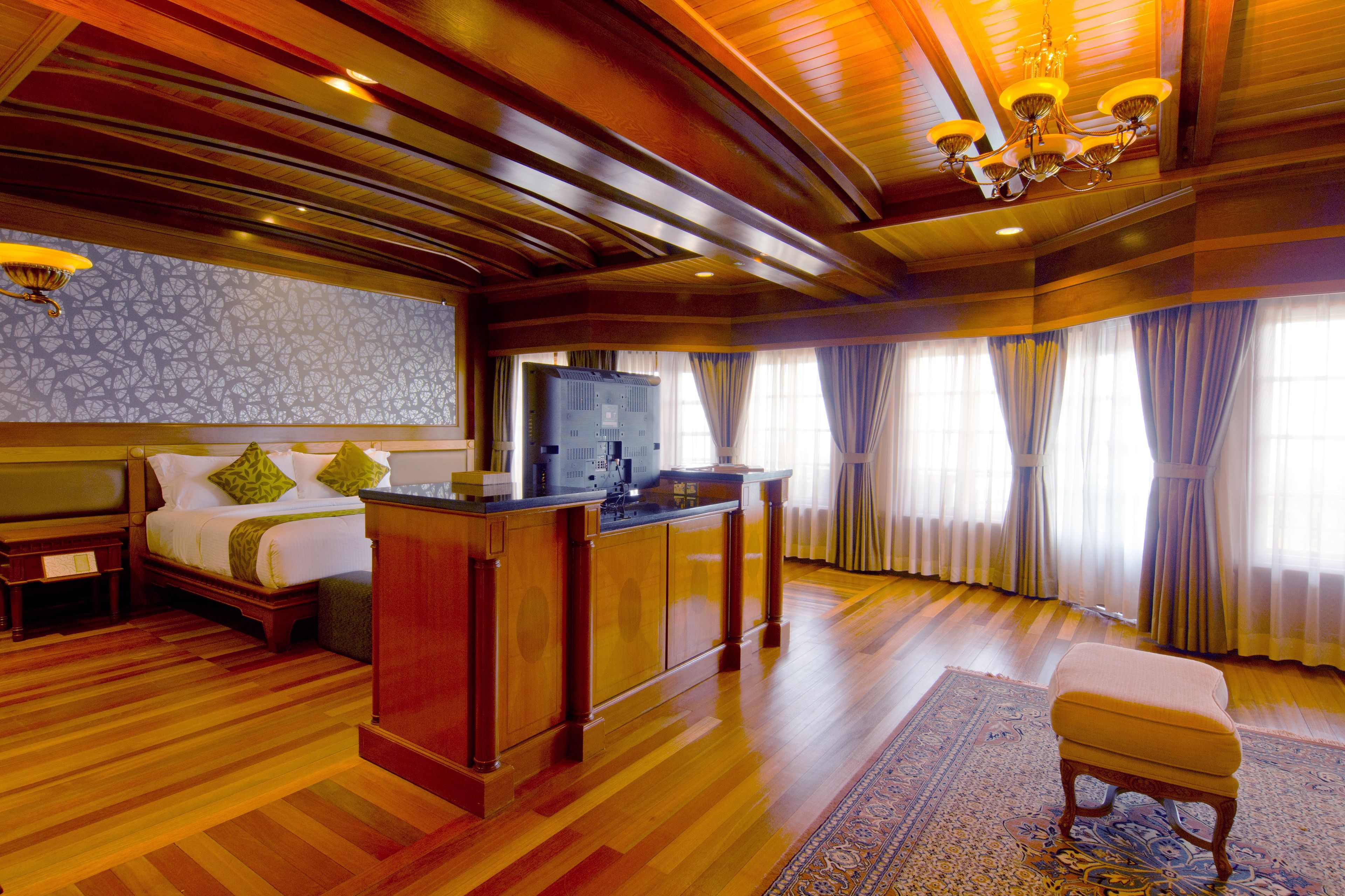 A hotel room with a wooden floor, a bed with pillows, a desk, a television, and curtains.