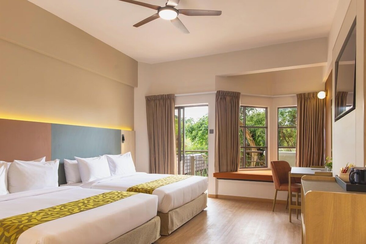A hotel room with two beds, a desk, a chair, a ceiling fan, and large windows with curtains.