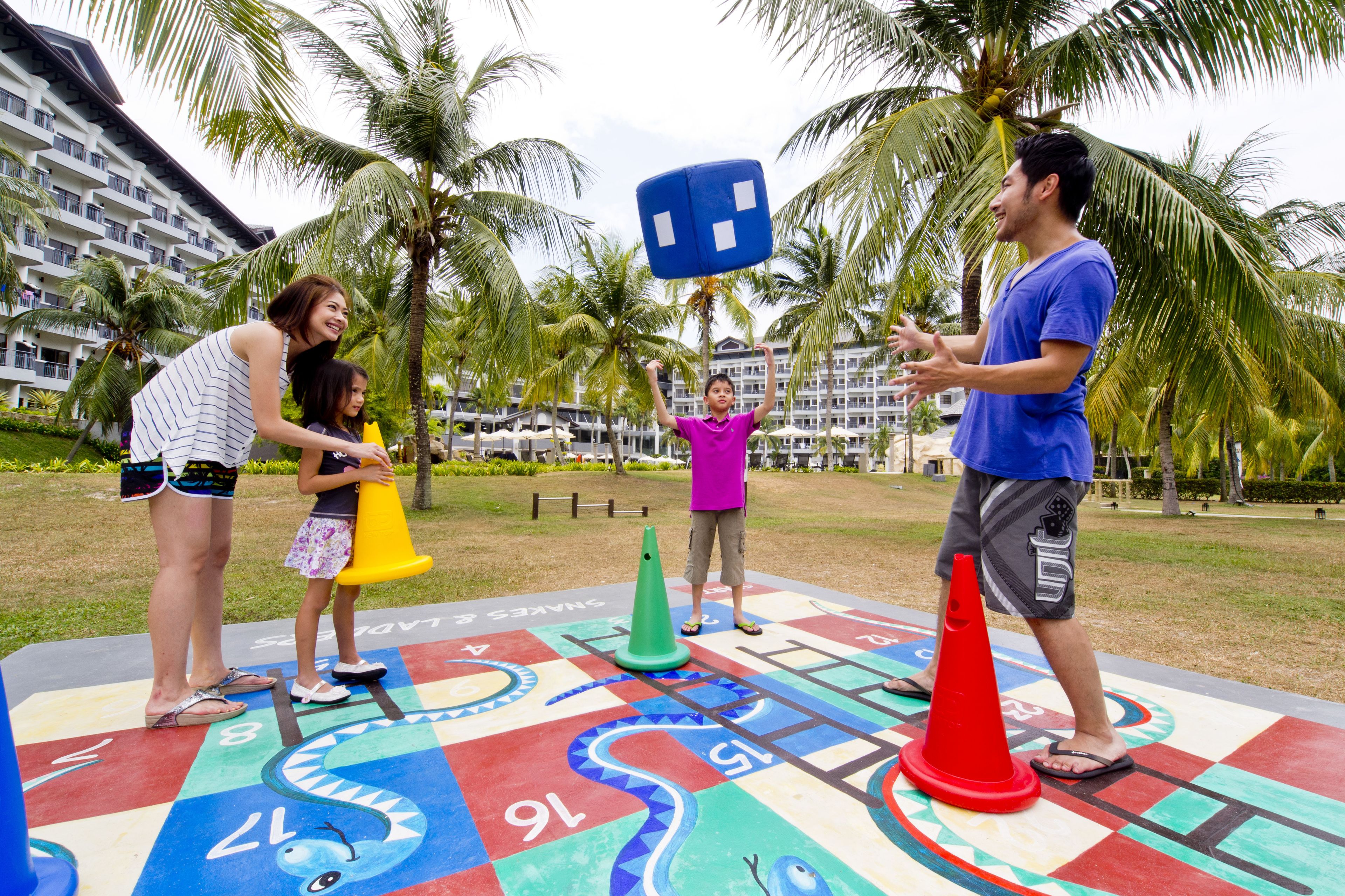 Un grupo de personas jugando un juego de mesa al aire libre, con palmeras y un hotel al fondo.