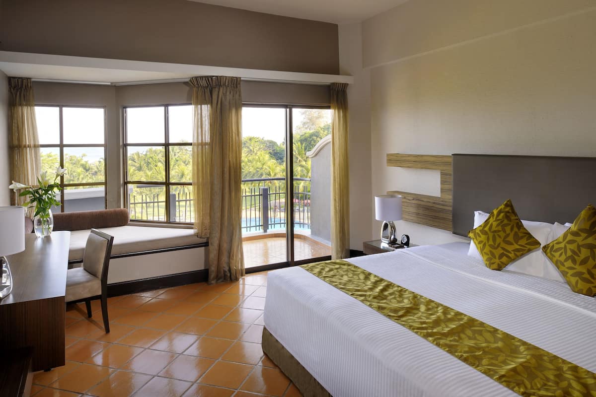 A hotel room with a large bed, a desk with a lamp, a chair, and a view of a balcony with a pool.