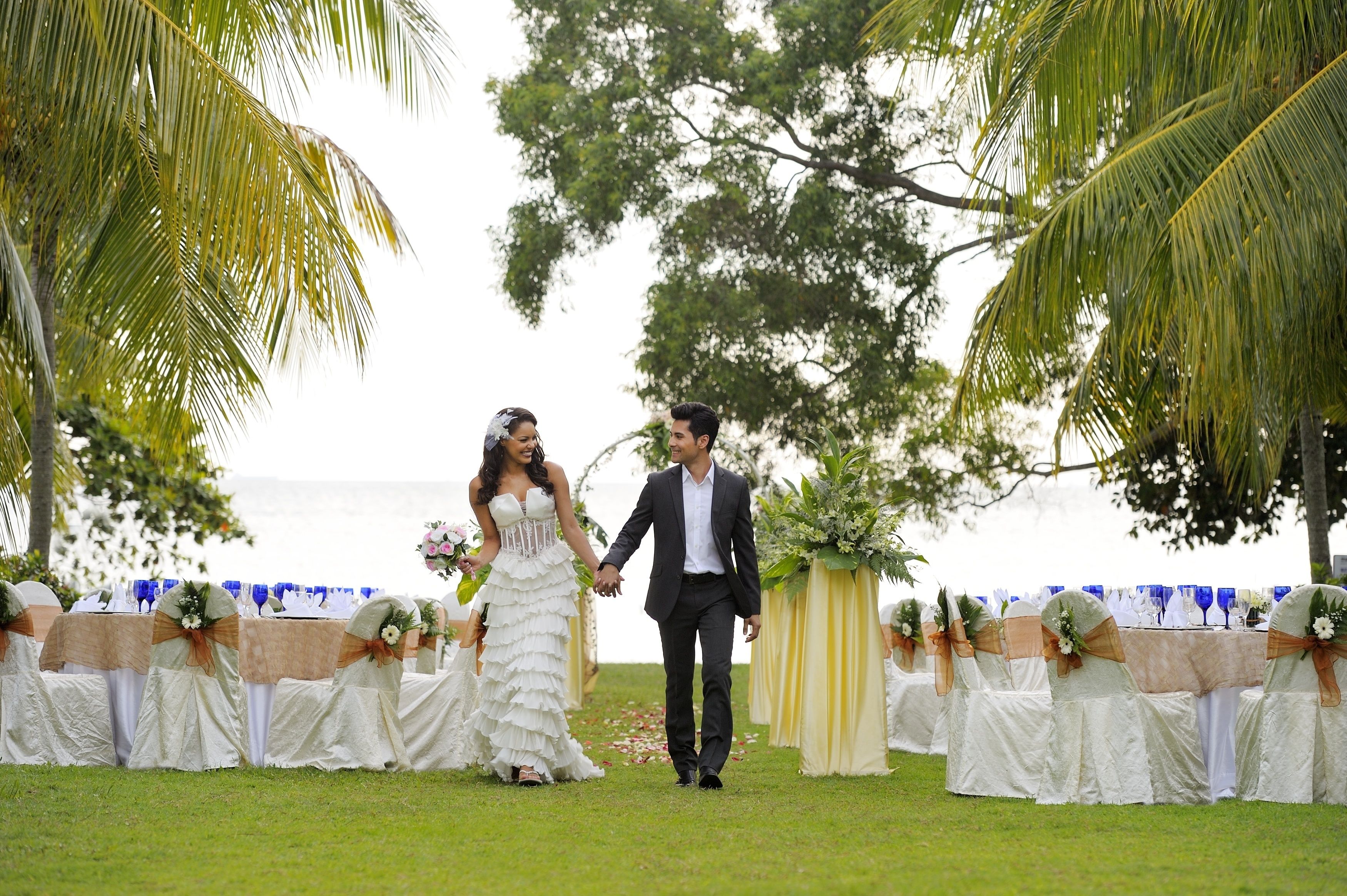 A wedding ceremony with a couple holding hands, surrounded by guests seated at tables with white tablecloths and floral centerpieces.