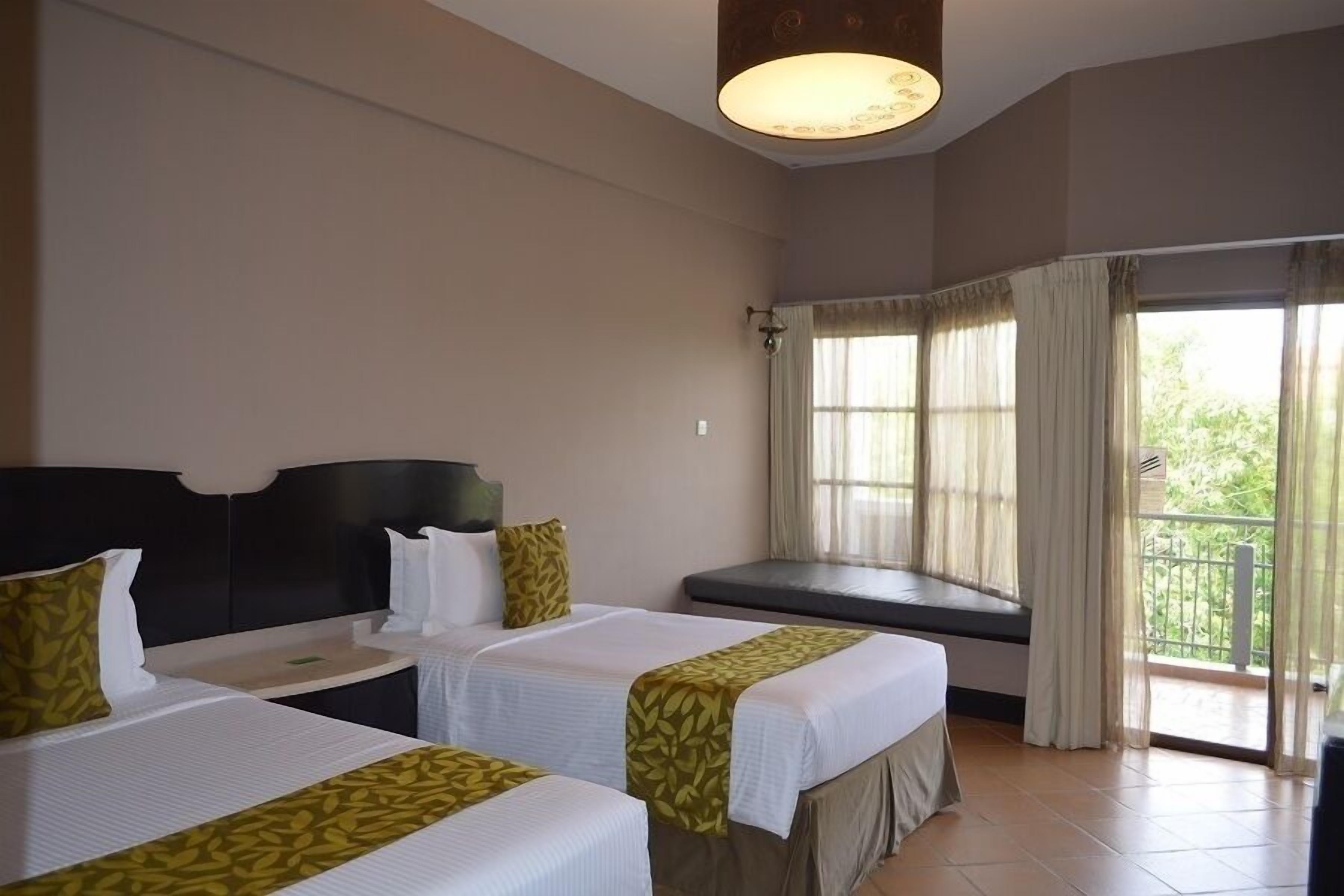 Quarto de hotel com duas camas, janela com cortinas, varanda e uma luminária no teto.