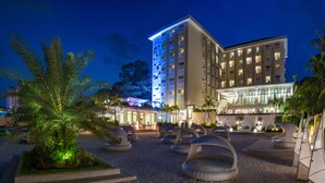 A hotel with a modern design, featuring a large building with multiple floors and a well-lit entrance. There are outdoor seating areas with unique, curved chairs and a landscaped garden with palm trees. - Be Resorts - Mactan (Lapu-Lapu)