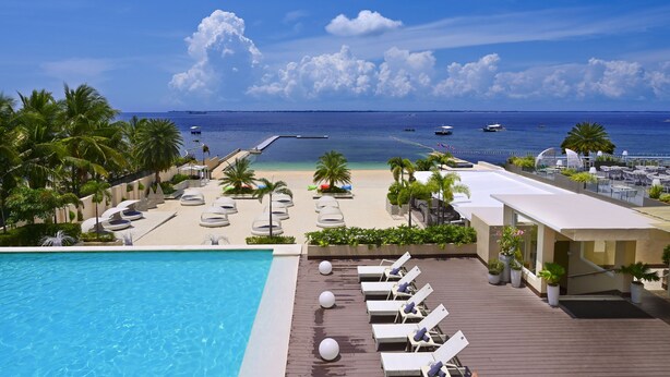 A resort pool area with lounge chairs, a beach view, and a clear sky. - Be Resorts - Mactan (Lapu-Lapu)