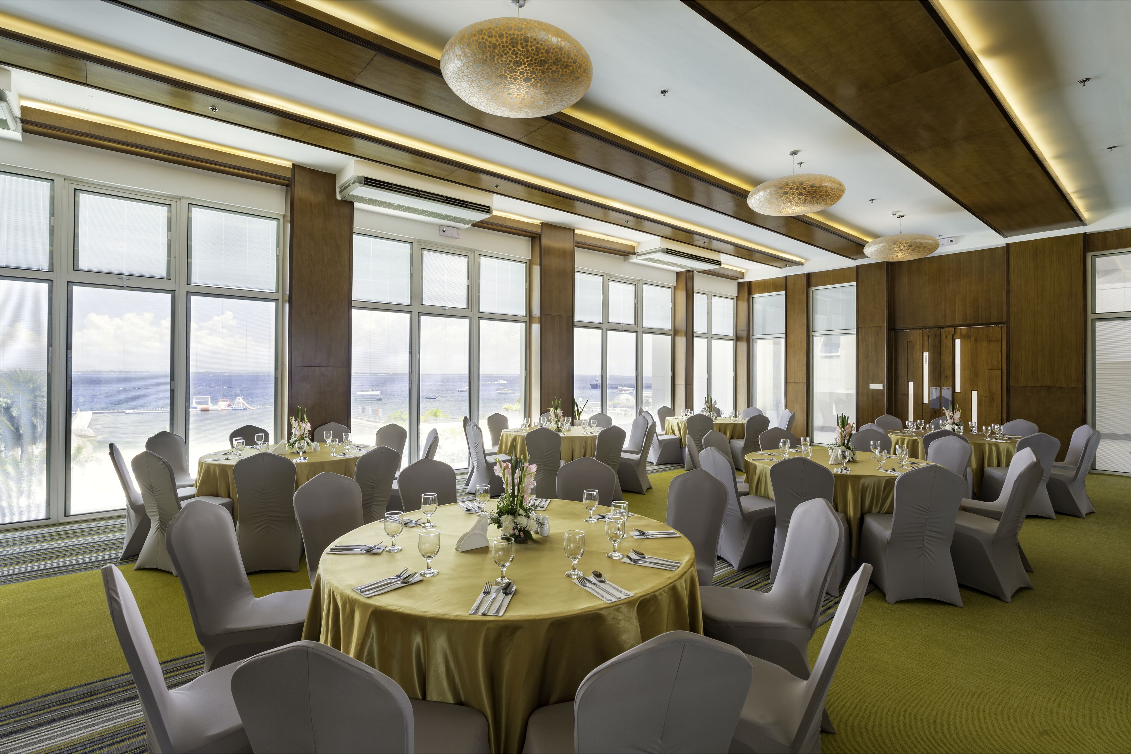 A banquet hall with round tables set for dining, large windows offering a view, and modern lighting fixtures.