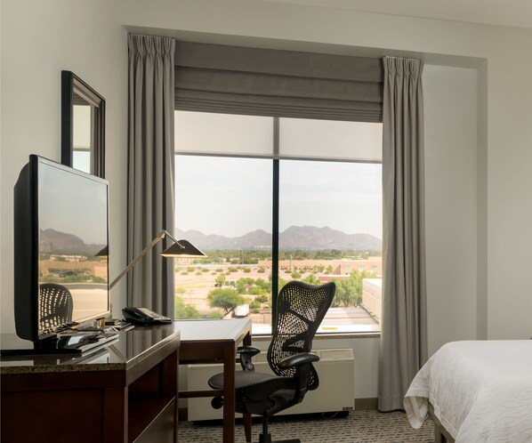 A hotel room with a desk, chair, bed, and a large window with curtains. - Hilton Garden Inn Phoenix Airport North (Phoenix)