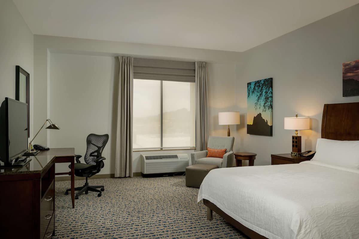 A hotel room with a bed, desk, chair, air conditioning unit, and two lamps.