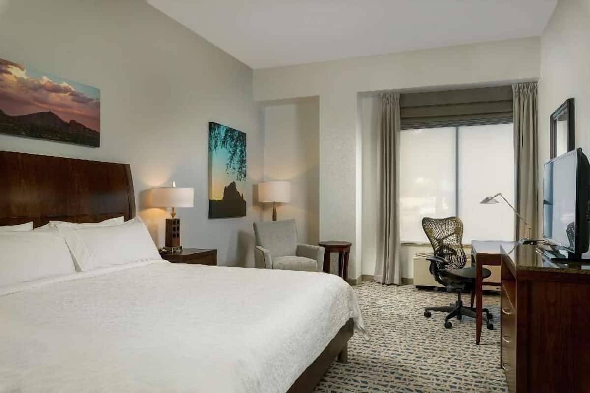 A hotel room with a large bed, two chairs, a desk, a television, and a window with curtains.