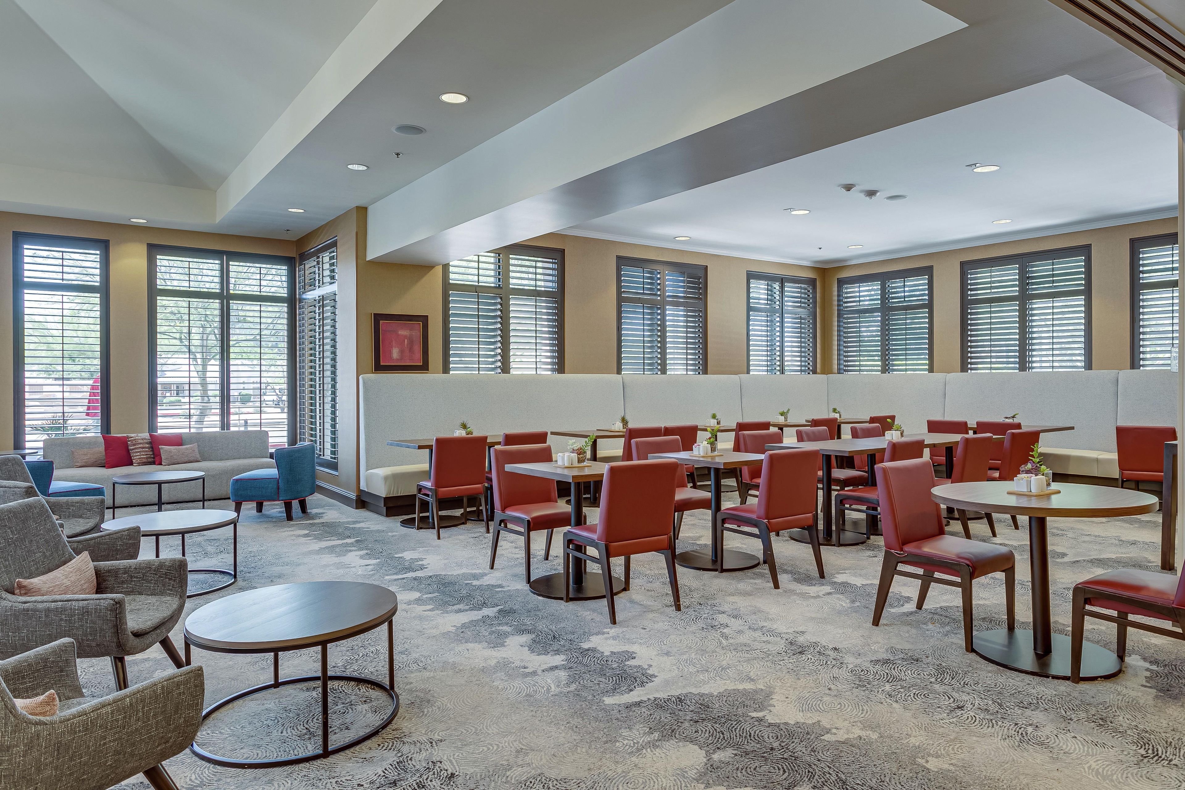 Une salle à manger d’hôtel avec de nombreuses tables et chaises, de grandes fenêtres équipées de stores et un tapis à motifs.