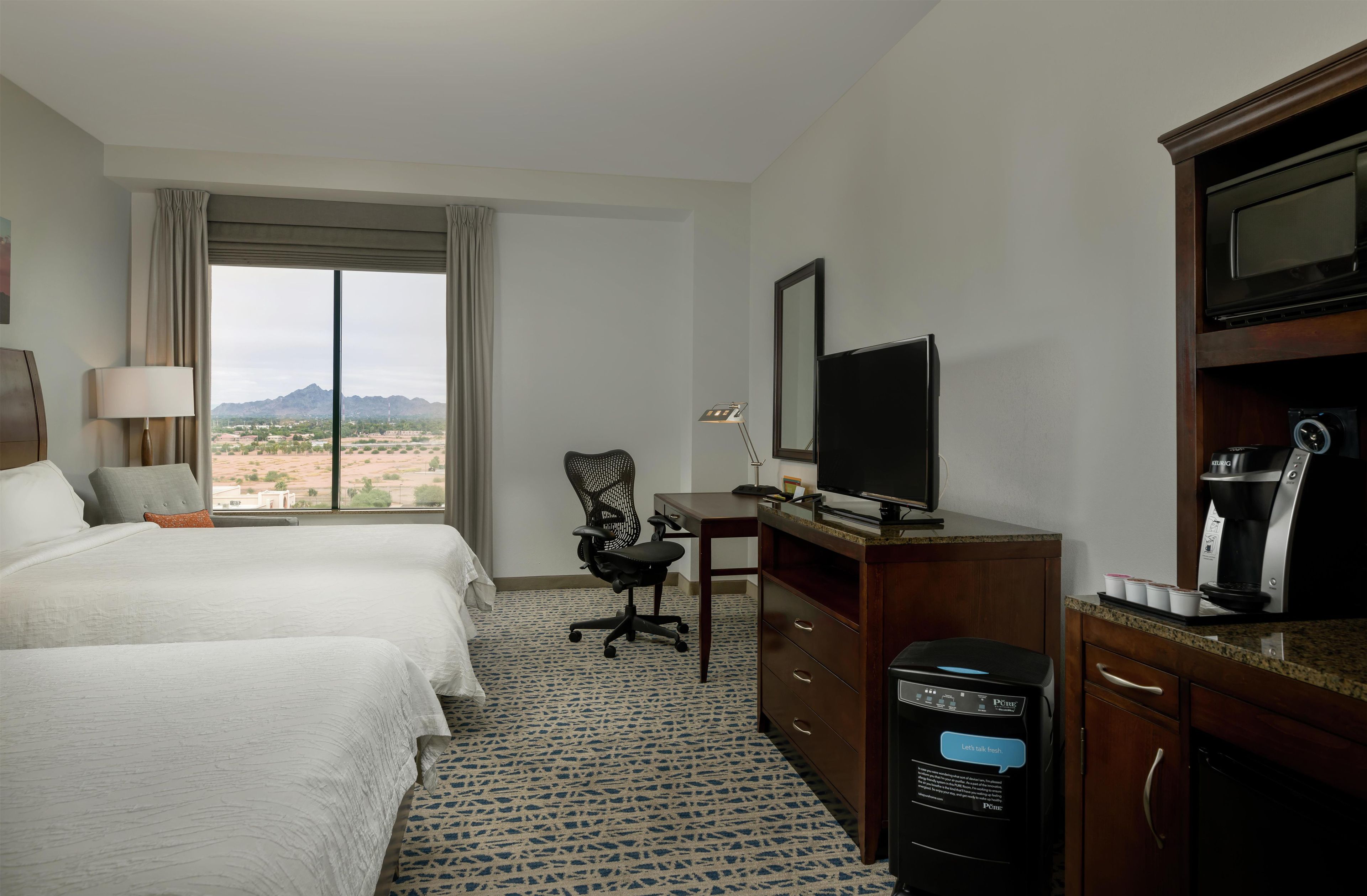 Habitación de hotel con dos camas, un escritorio con televisor, cafetera y vistas a un paisaje desértico.