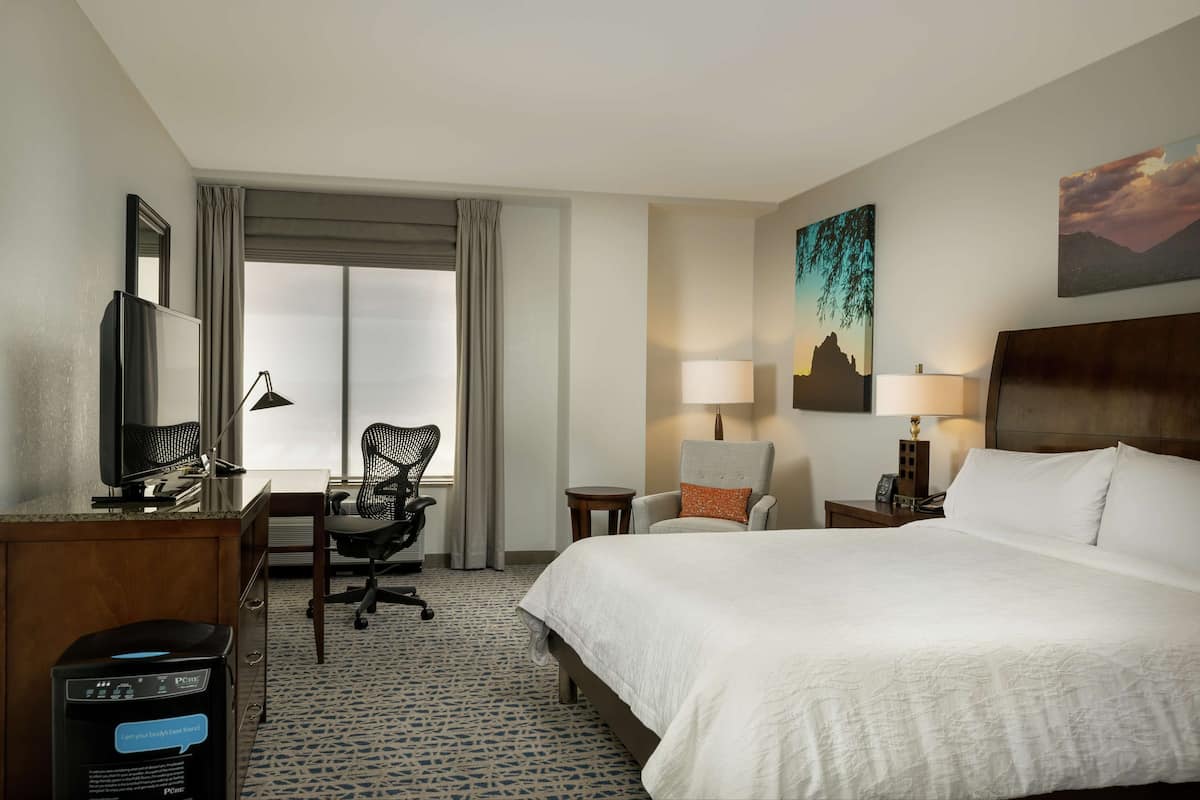 A hotel room with a large bed, a desk with a computer, a chair, a TV, and a window with curtains.