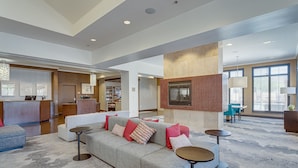 A spacious living area with a sofa, ottoman, and round tables, a kitchen in the background, and large windows providing natural light. - Hilton Garden Inn Phoenix Airport North (Phoenix)