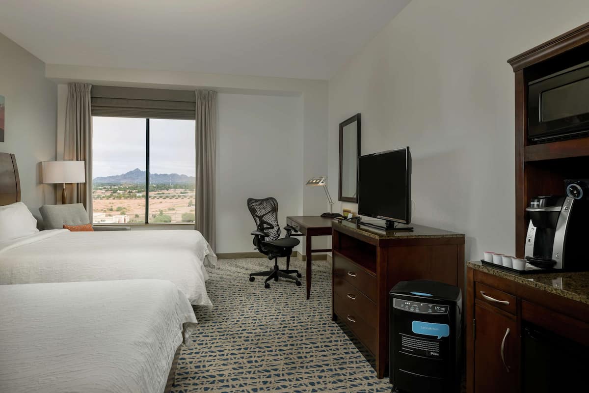 A hotel room with two beds, a desk with a television, a coffee maker, and a view of a desert landscape.