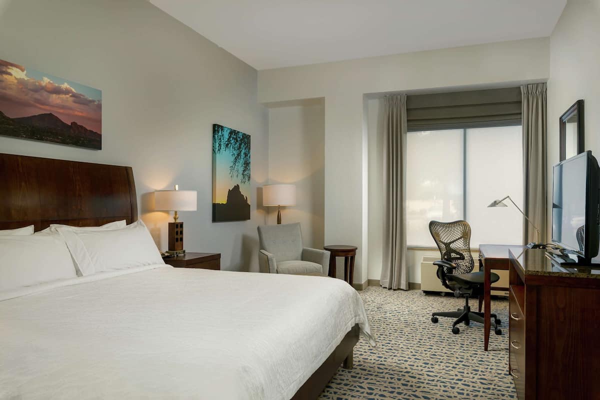 A hotel room with a large bed, two chairs, a desk, a TV, and a window with curtains.