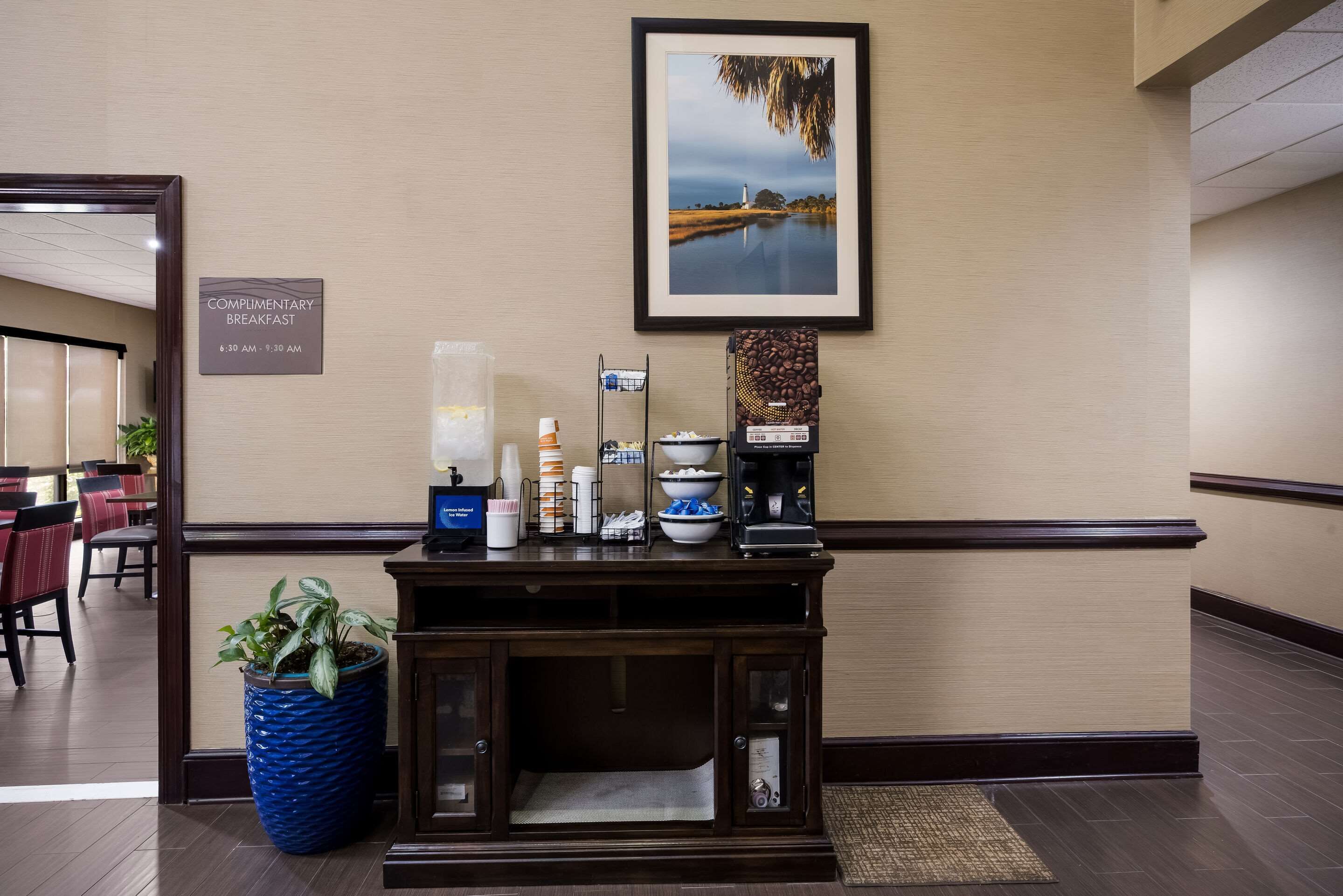 A hotel reception area with a complimentary breakfast sign, a coffee machine, a framed landscape photo, and a potted plant.