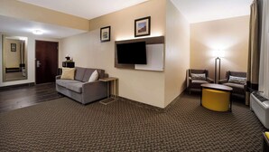 A hotel room with a sofa, a small table, a flat-screen TV mounted on the wall, a standing lamp, and a carpeted floor. - Comfort Inn & Suites Midway - Tallahassee West (Midway)