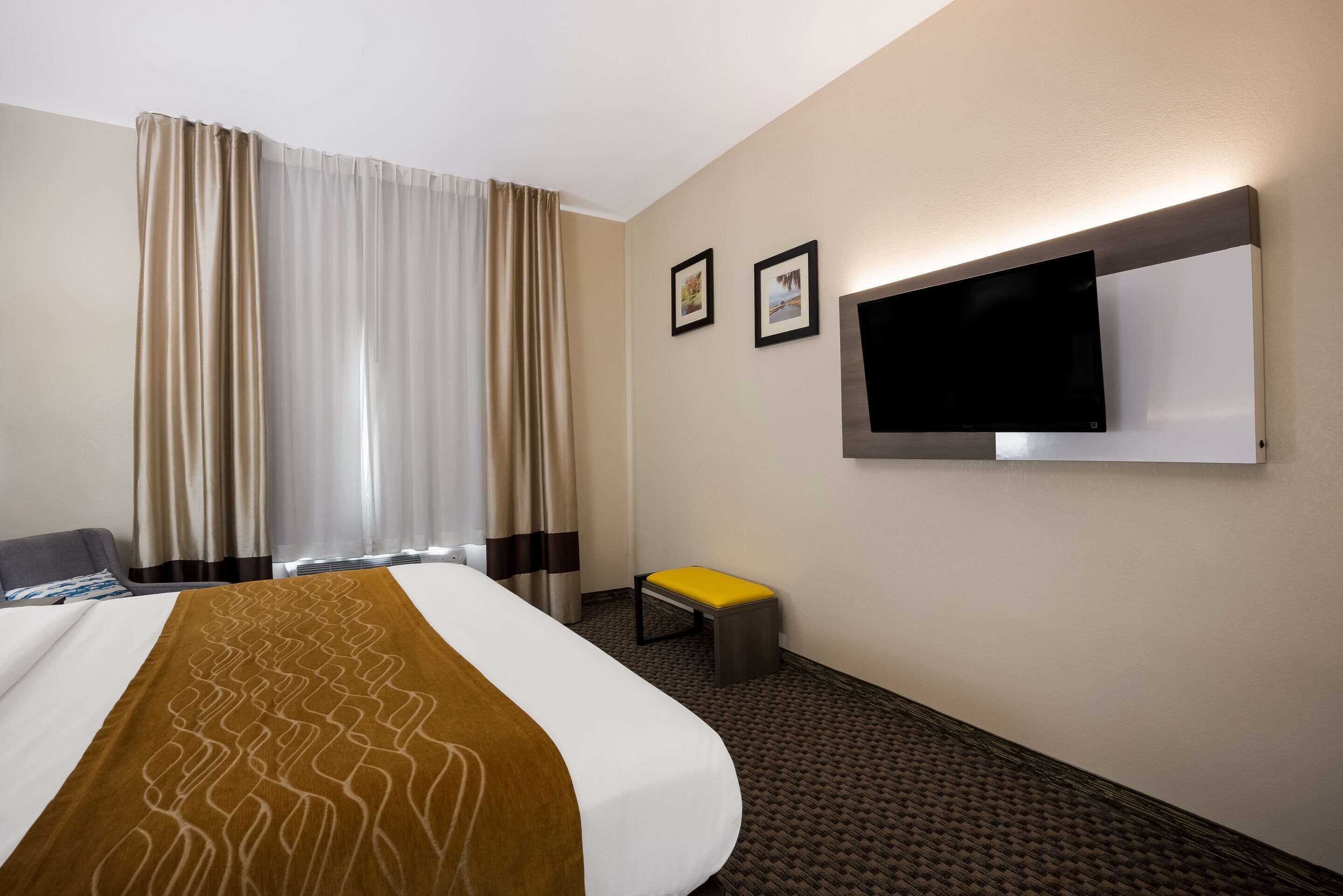 Ein Hotelzimmer mit einem Bett, einem an der Wand befestigten Fernseher, einem kleinen Hocker und Vorhängen.