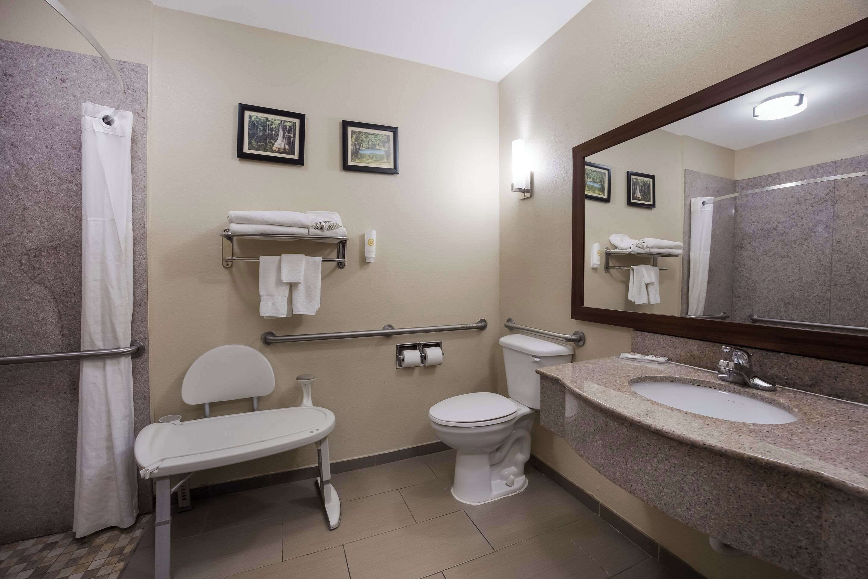 Un bany amb un vàter blanc, un lavabo, una cadira, una dutxa i dos quadres amb marc a la paret.