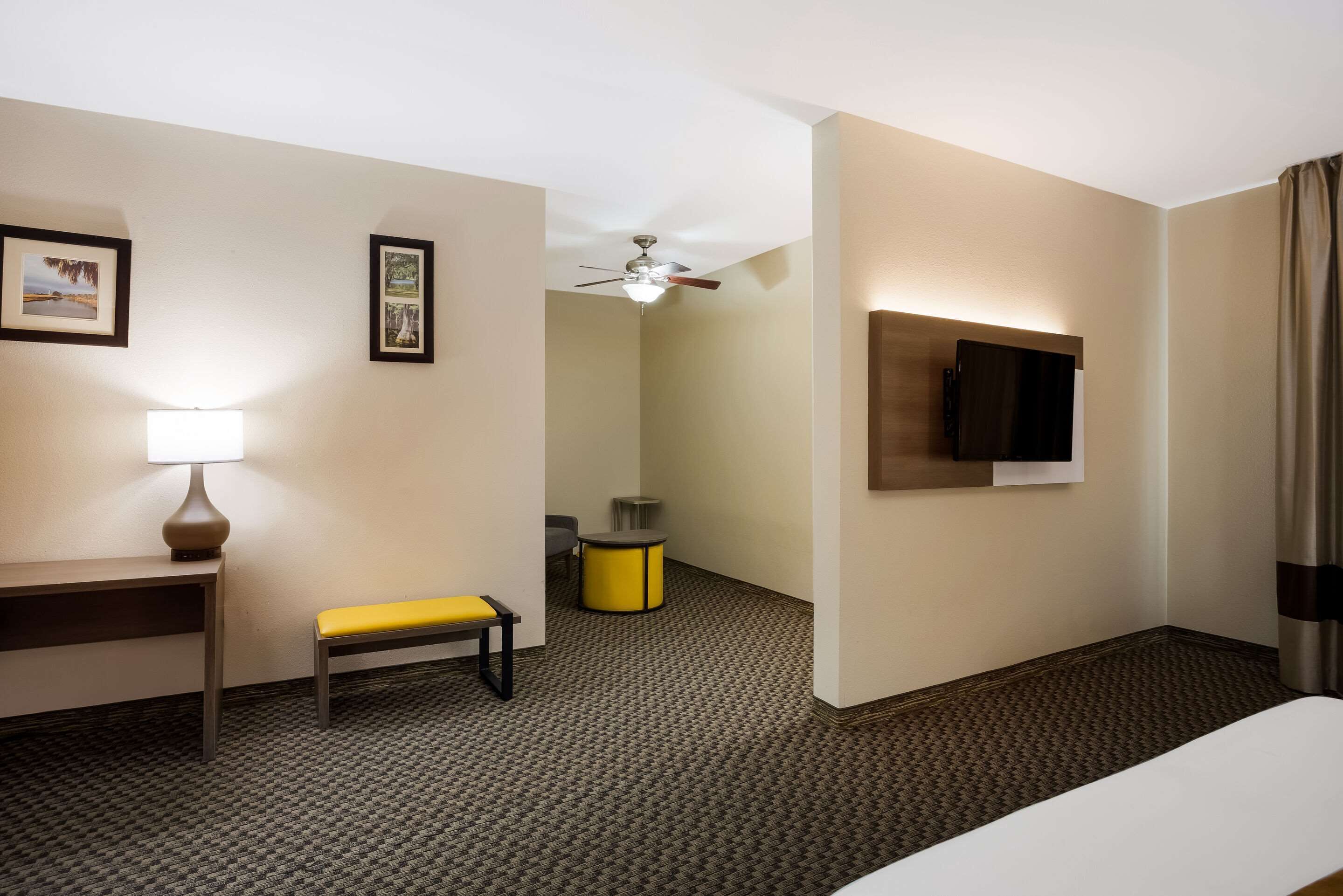 Habitación de hotel con cama, escritorio con lámpara, banco, ventilador de techo, televisor montado en la pared y una pequeña mesa con una silla.