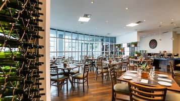A spacious restaurant with high ceilings, large windows, and wooden flooring. Tables are set with white plates and glasses, and a wine rack is visible on the left.